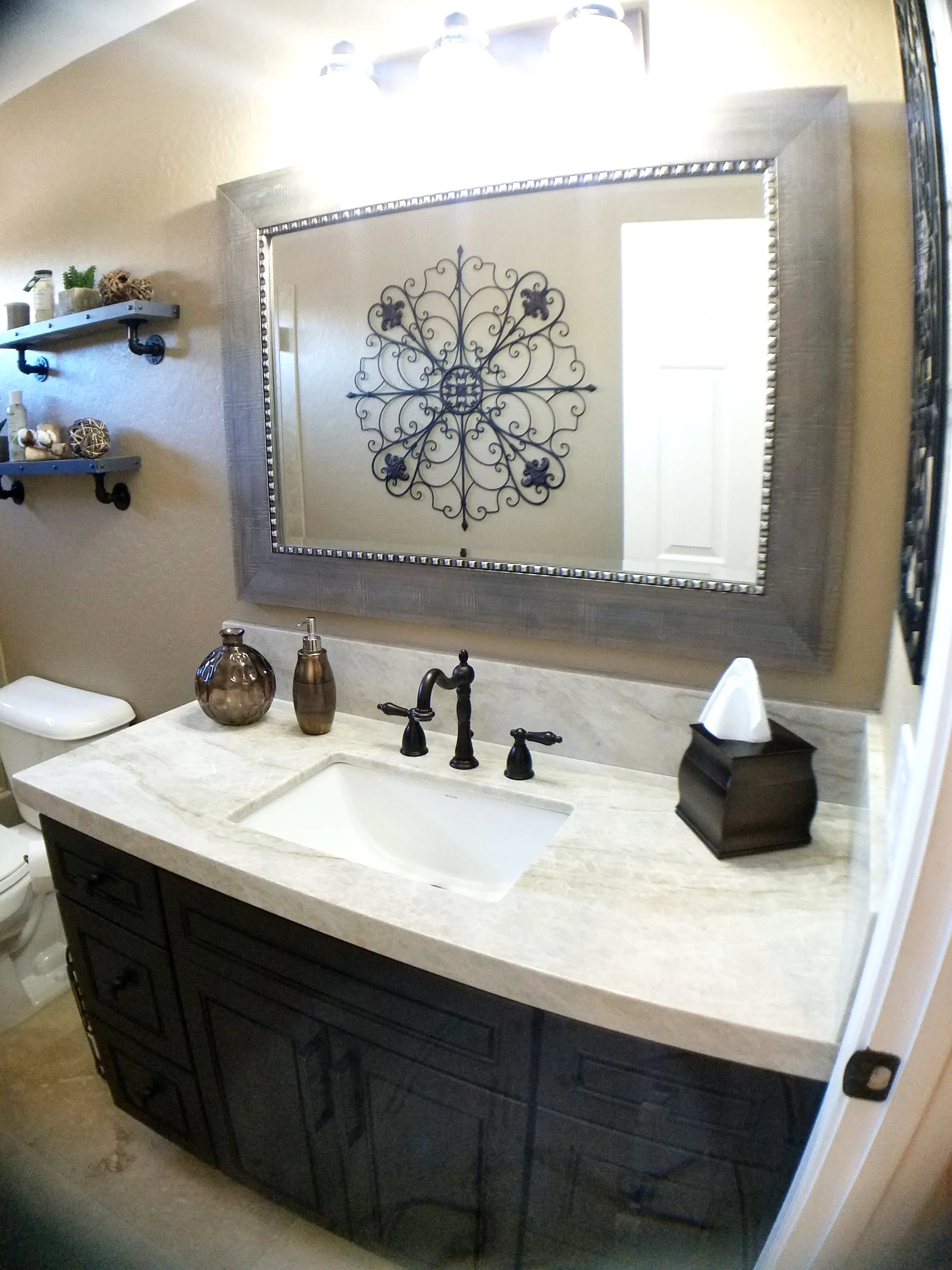 Dark carved bathroom vanity with a pale stone top, framed mirror and industrial pipe shelving on a textured wall